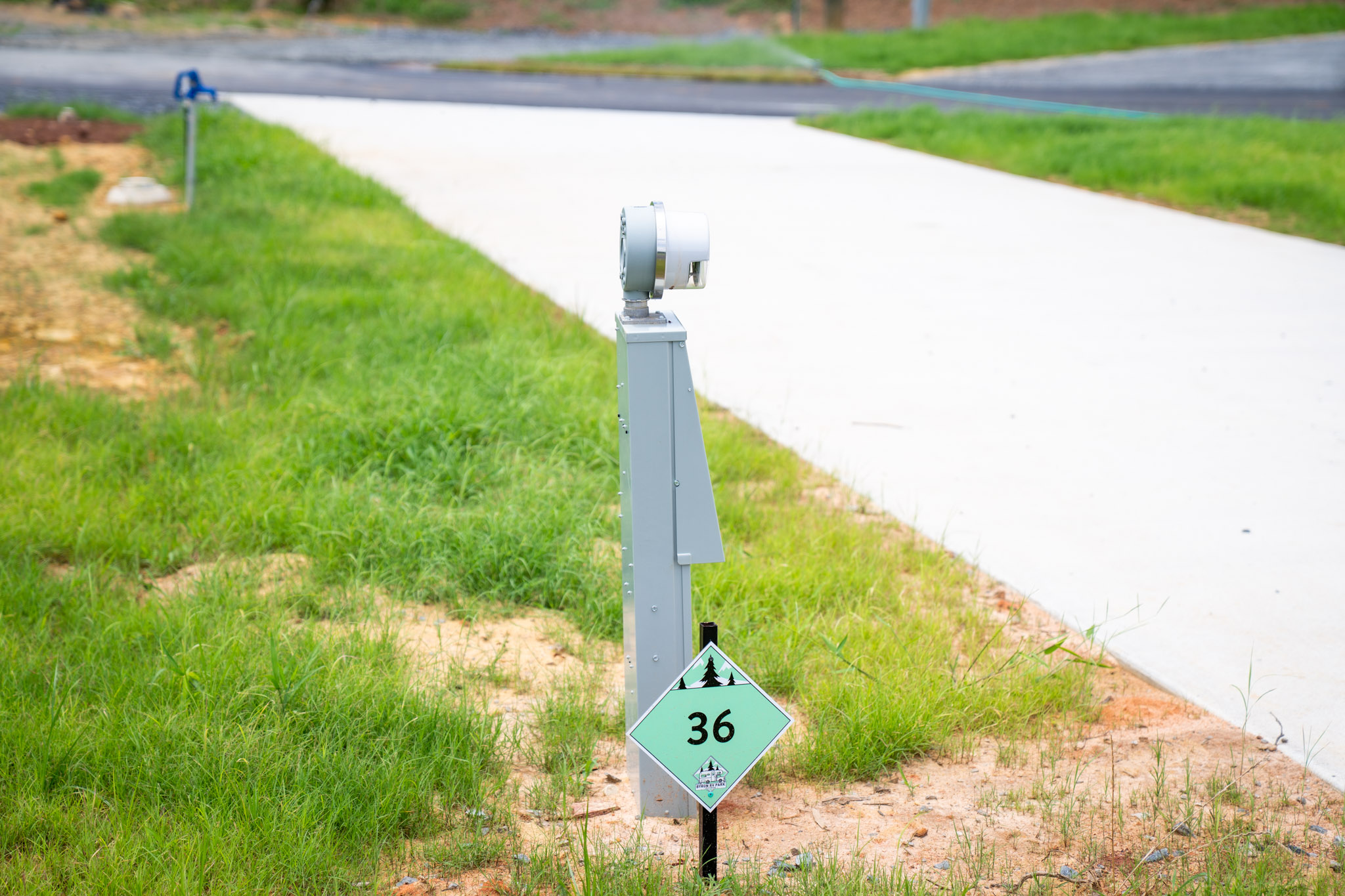 A close-up of an electrical hookup post at an RV site with a green site marker labeled “36” on grass beside a paved driveway.