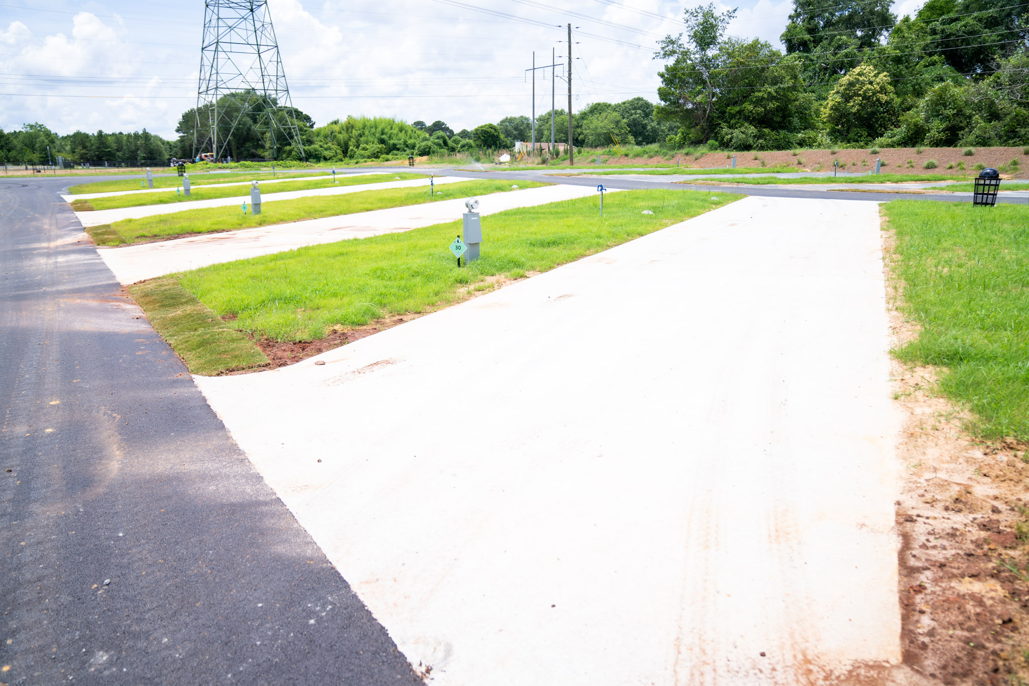A row of empty concrete RV pads with green grass strips and utility hookups, under a partly cloudy sky, in a newly developed RV park. Trees and power lines are visible in the background.