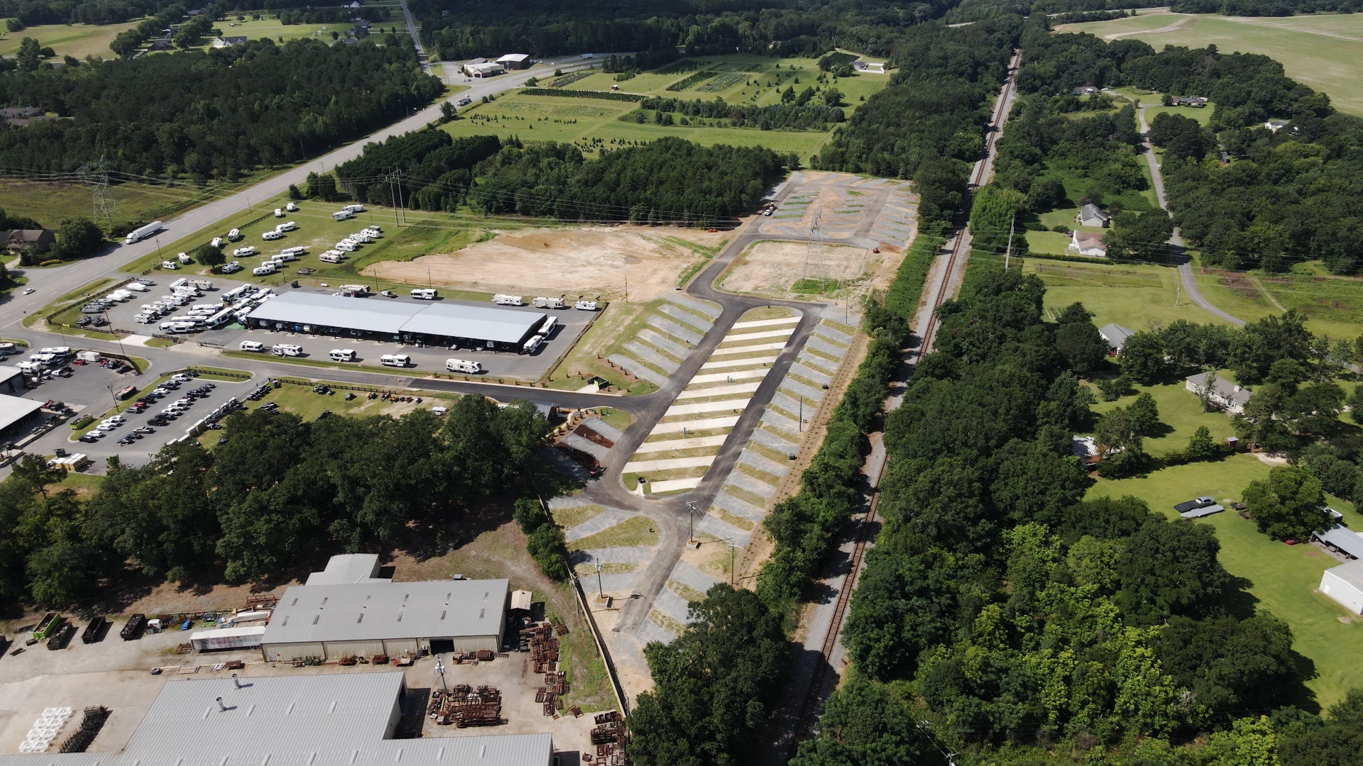 Aerial view of a construction site with paved roads and marked lots surrounded by trees, buildings, a train track to the right, and parked vehicles to the left on a sunny day.