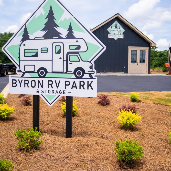 A large sign reading "Byron RV Park & Storage" stands in front of a dark building with matching logo. The area is landscaped with mulch and green shrubs under a partly cloudy sky.