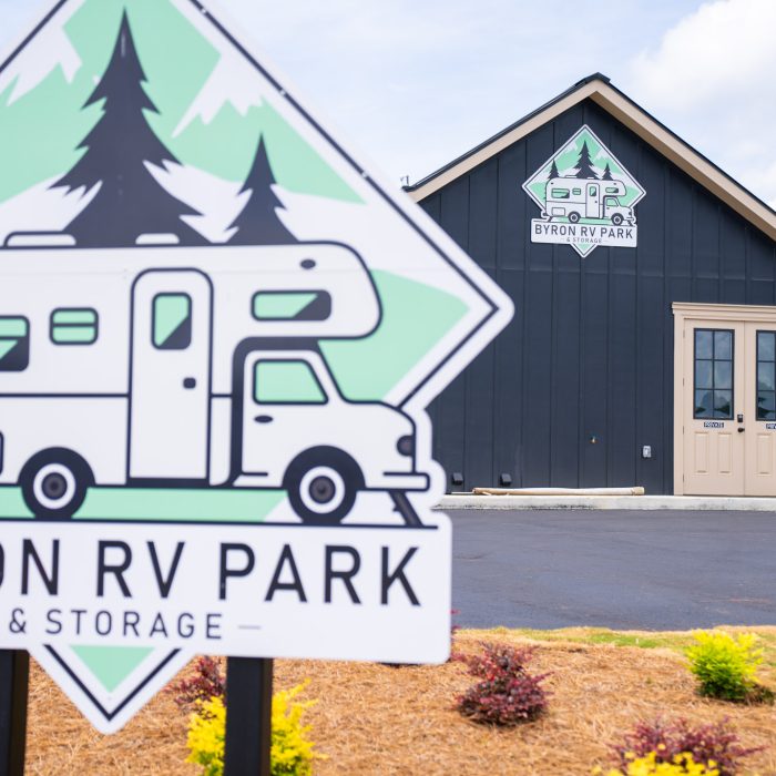 Sign for Byron RV Park & Storage in the foreground, featuring an RV and pine trees. In the background is a dark building with double doors and a matching Byron RV Park sign above the entrance.