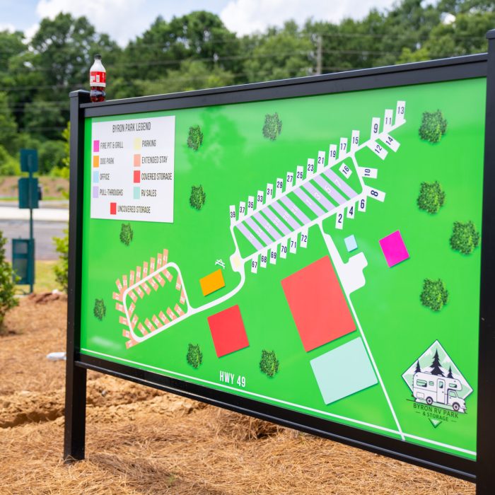 A colorful map sign for a campground or RV park stands outdoors by a road, showing numbered lots, color-coded areas, trees, and the label "HWY 49." Green landscape and trees are visible in the background.