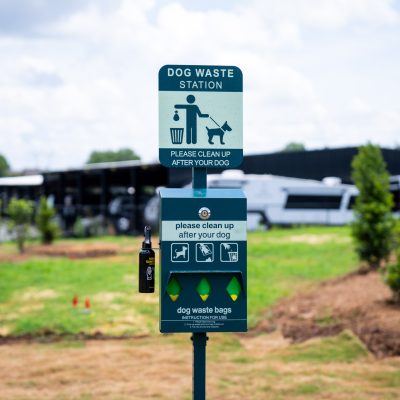 A green dog waste station with a sign reminding owners to clean up after their dogs, located in a grassy area with trees and parked vehicles in the background.