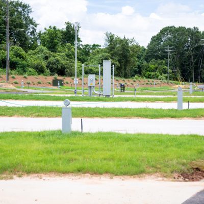 Several empty RV parking spots with utility hookups on concrete pads, surrounded by green grass and trees in the background, under a partly cloudy sky.