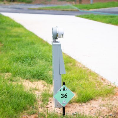 A gray electrical hookup post stands beside a paved driveway, with a green diamond-shaped sign marked “36” and small patches of grass and dirt surrounding the area.