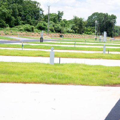 Rows of empty concrete RV pads with utility hookups are set in a grassy area, surrounded by trees and utility poles under a partly cloudy sky.
