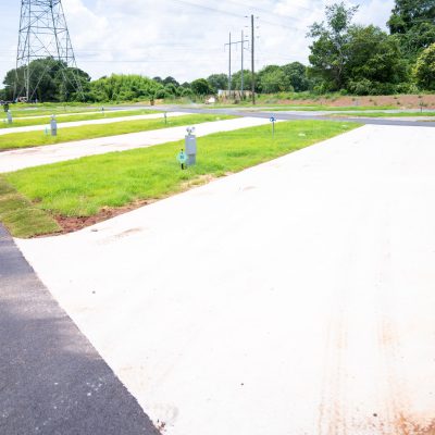 A row of empty concrete RV pads with green grass strips and utility hookups, under a partly cloudy sky, in a newly developed RV park. Trees and power lines are visible in the background.