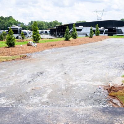 A gravel RV parking pad with young trees on the left, bordered by grass and a water hookup on the right. Several RVs and storage buildings are visible in the background under a partly cloudy sky.
